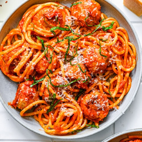An overhead shot of a white plate of chicken meatballs tossed with sauce and noodles on a white background
