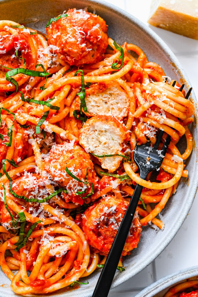 A white plate with chicken meatballs tossed in sauce and noodles with a fork on the plate with spaghetti twisted on it