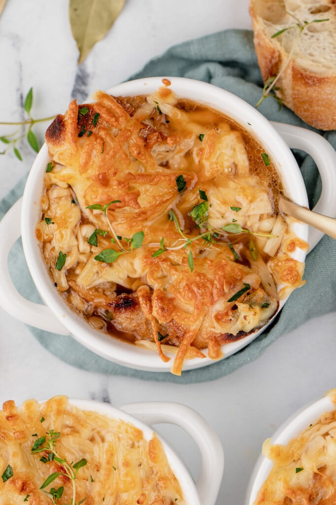 An overhead shot of a white bowl of easy french onion soup on a blue towel with another bowl and baguette on the side