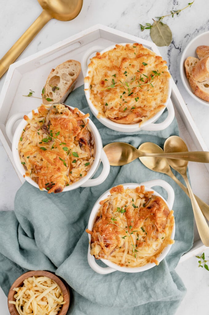 An overhead shot of three white bowls of healthy french onion soup on a blue towel with gold spoons on the side