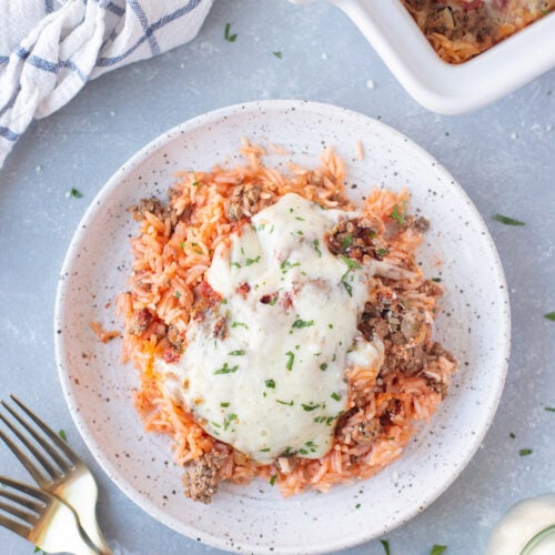 An overhead shot of a white plate with a large helping of hamburger casserole with a baking dish with casserole, a cloth, and forks on the side