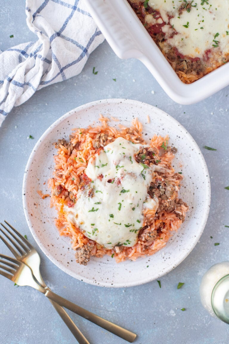 An overhead shot of a white plate with a large helping of hamburger casserole with a baking dish with casserole, a cloth, and forks on the side