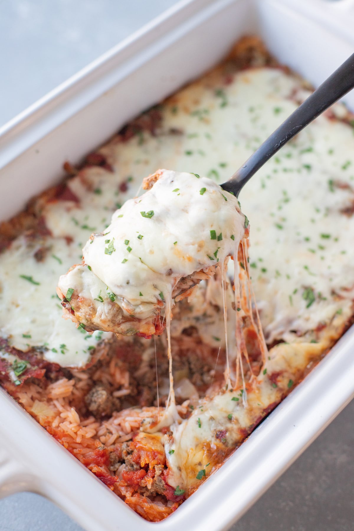 A white baking dish of hamburger casserole with a spoon taking a large scoop of the casserole with a cheese pull