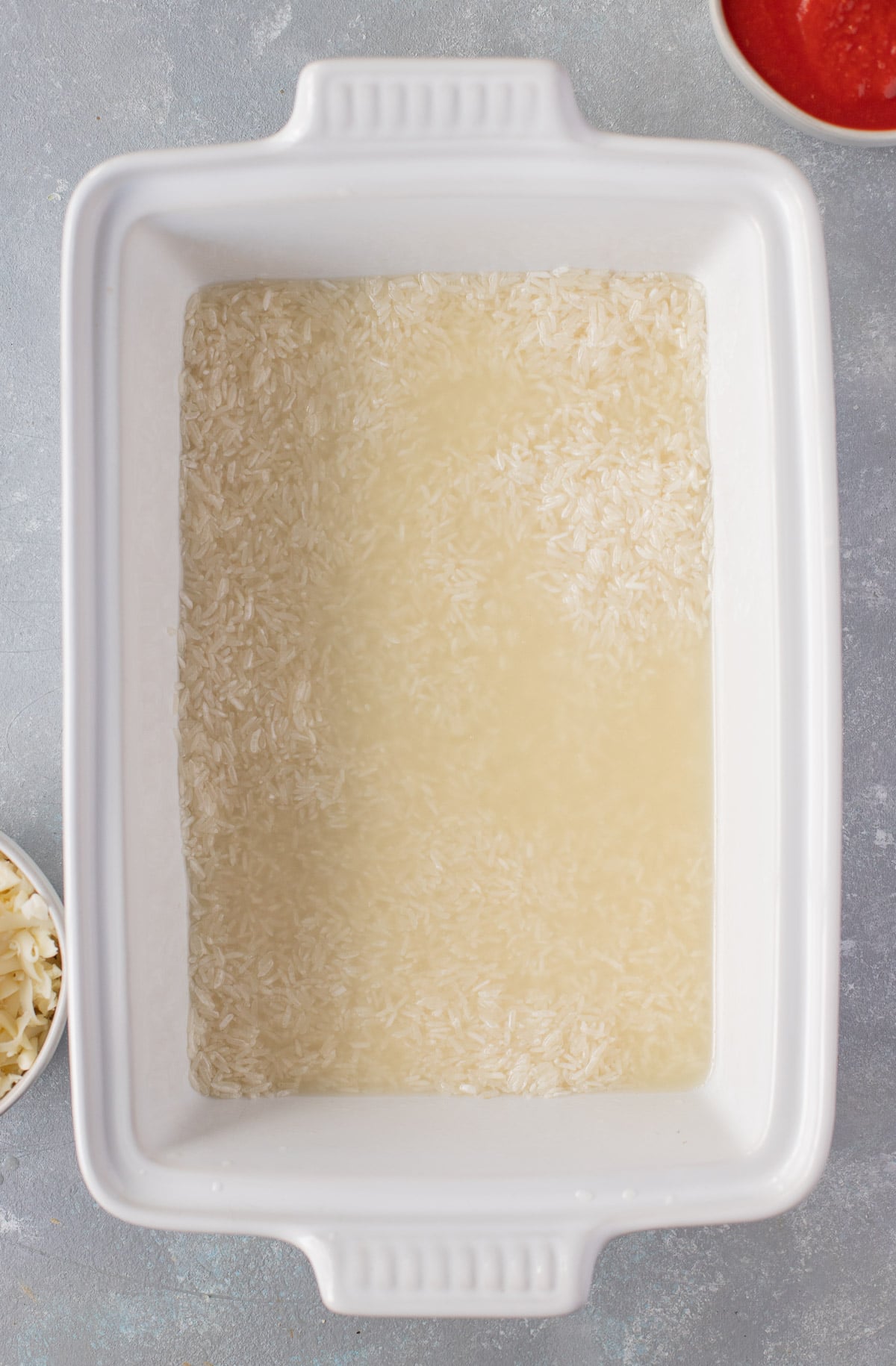 An overhead shot of a white baking dish with rice and chicken broth for hamburger casserole