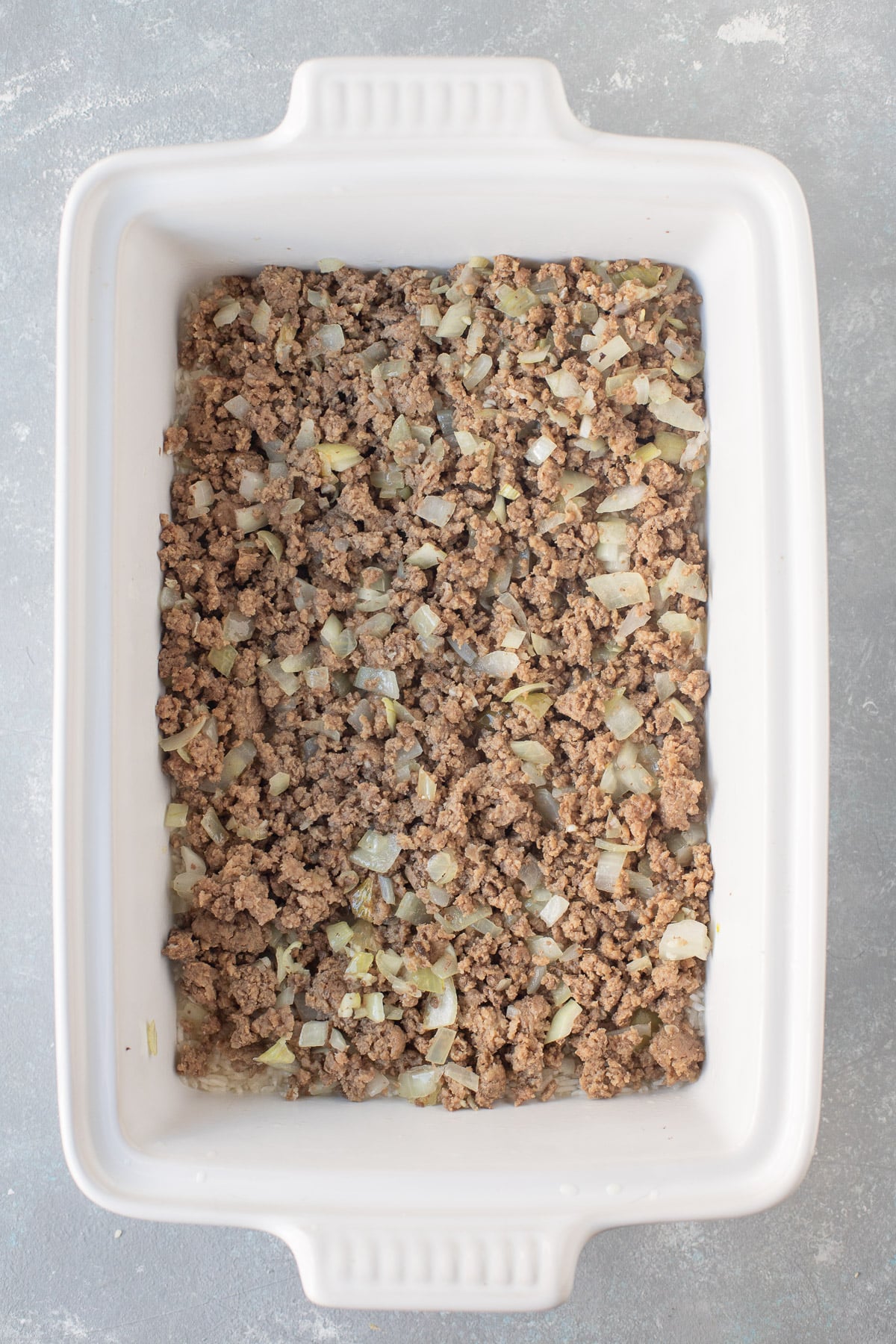 An overhead shot of a white baking dish with ground beef layered on top of the rice and chicken broth for hamburger casserole