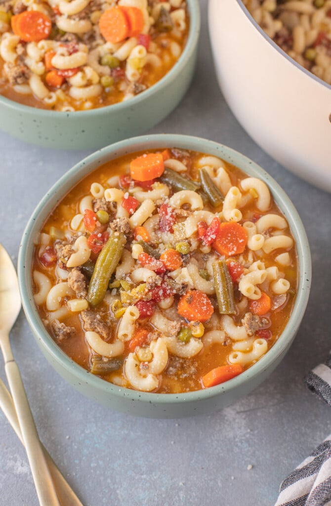 A bowl of hamburger vegetable soup on a grey background with gold spoons on the side and another bowl in the background