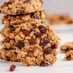 A oatmeal raisin cookie leaning on a stack of cookies on a grey background