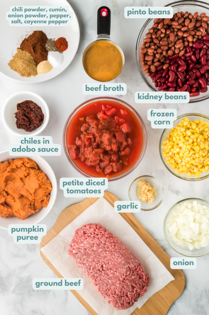 Ingredients for pumpkin chili in white bowls: onion, garlic, ground beef, diced tomatoes, pumpkin puree, whole corn kernels, beef broth, pinto beans, kidney beans, chiles in adobo sauce, olive oil, chili powder, cumin, onion powder, salt, pepper, cayenne pepper