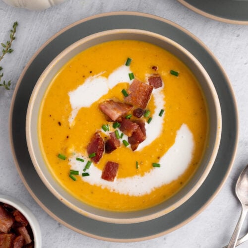 An overhead shot of roasted butternut squash soup in a grey bowl on a grey background with bacon and chives on top