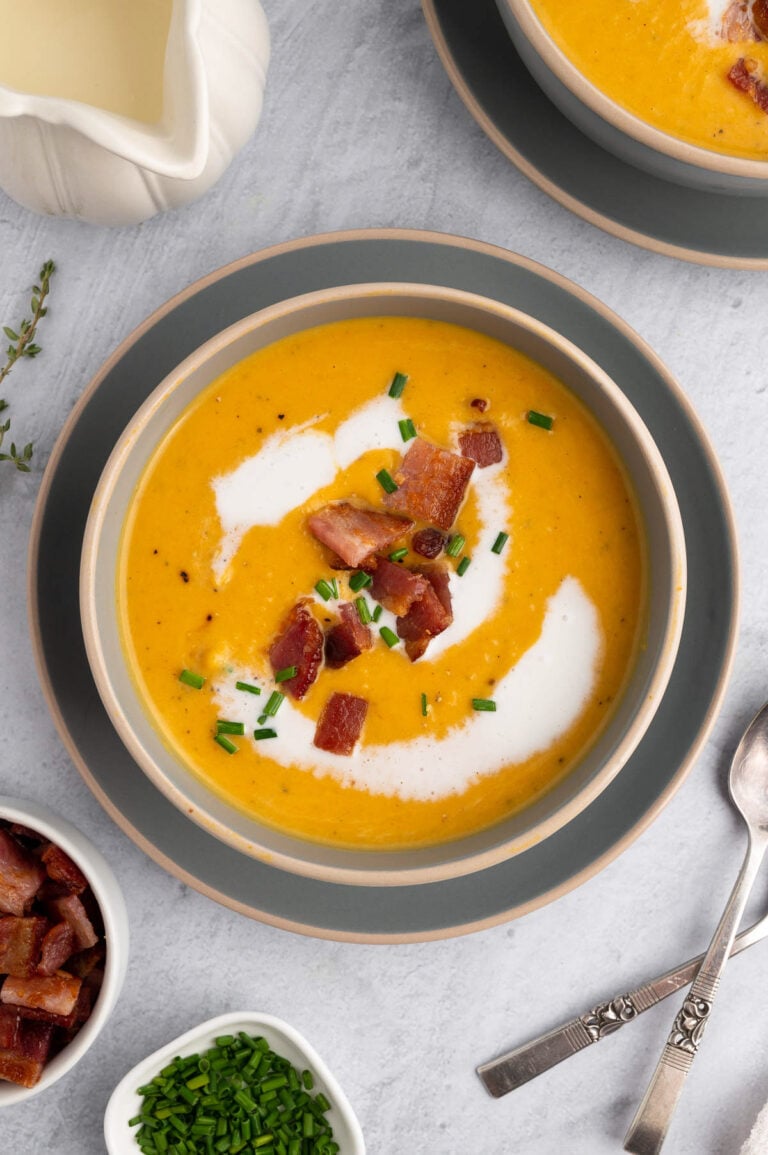 An overhead shot of roasted butternut squash soup in a grey bowl on a grey background with bacon and chives on top