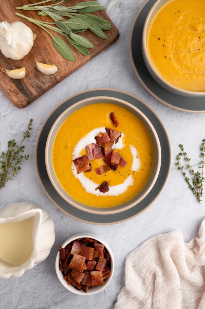 An overhead shot of roasted butternut squash soup in a grey bowl on a grey background with bacon and chives on top and another bowl of soup on the side