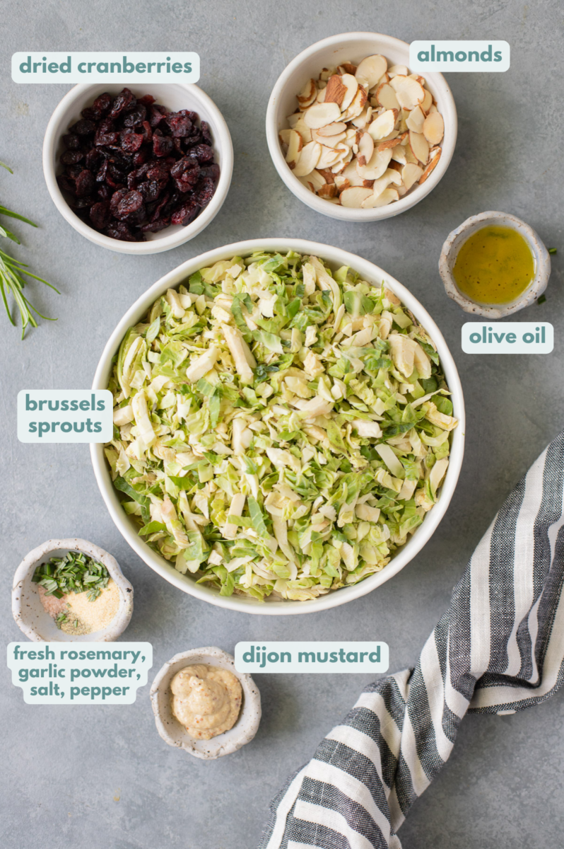 Shaved Brussel Sprout Salad Ingredients in white bowls - shaved brussels sprouts, almonds, dried cranberries, hemp seeds, olive oil, rosemary, salt, pepper, balsamic vinegar