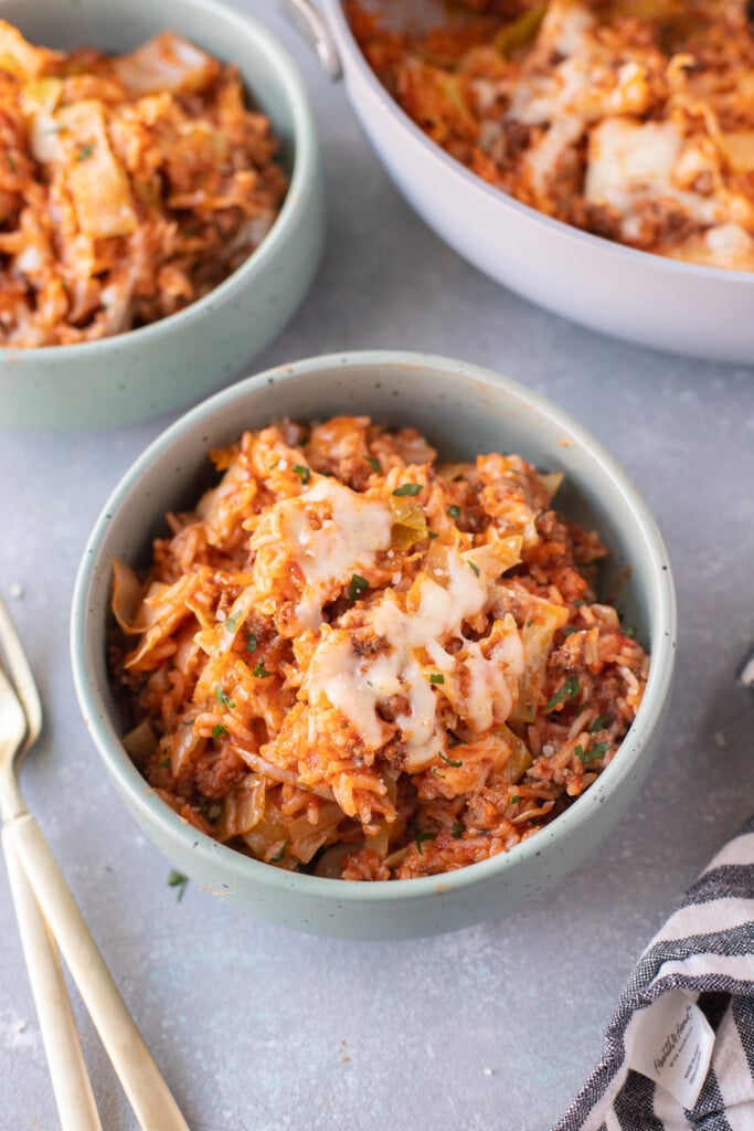 A bowl of unstuffed cabbage rolls recipe with another bowl, skillet, and forks on the side on a grey background
