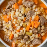 An overhead shot of a bowl of beef barley soup on a grey background with gold spoons on the side