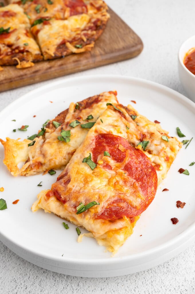 A white plate with two pieces of keto pizza with cauliflower pizza crust with a board of pizza in the background