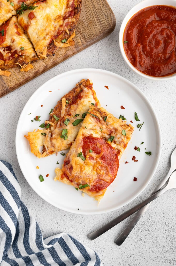 A white plate with two pieces of keto pizza with cauliflower pizza crust with a board of pizza and pizza sauce on the side