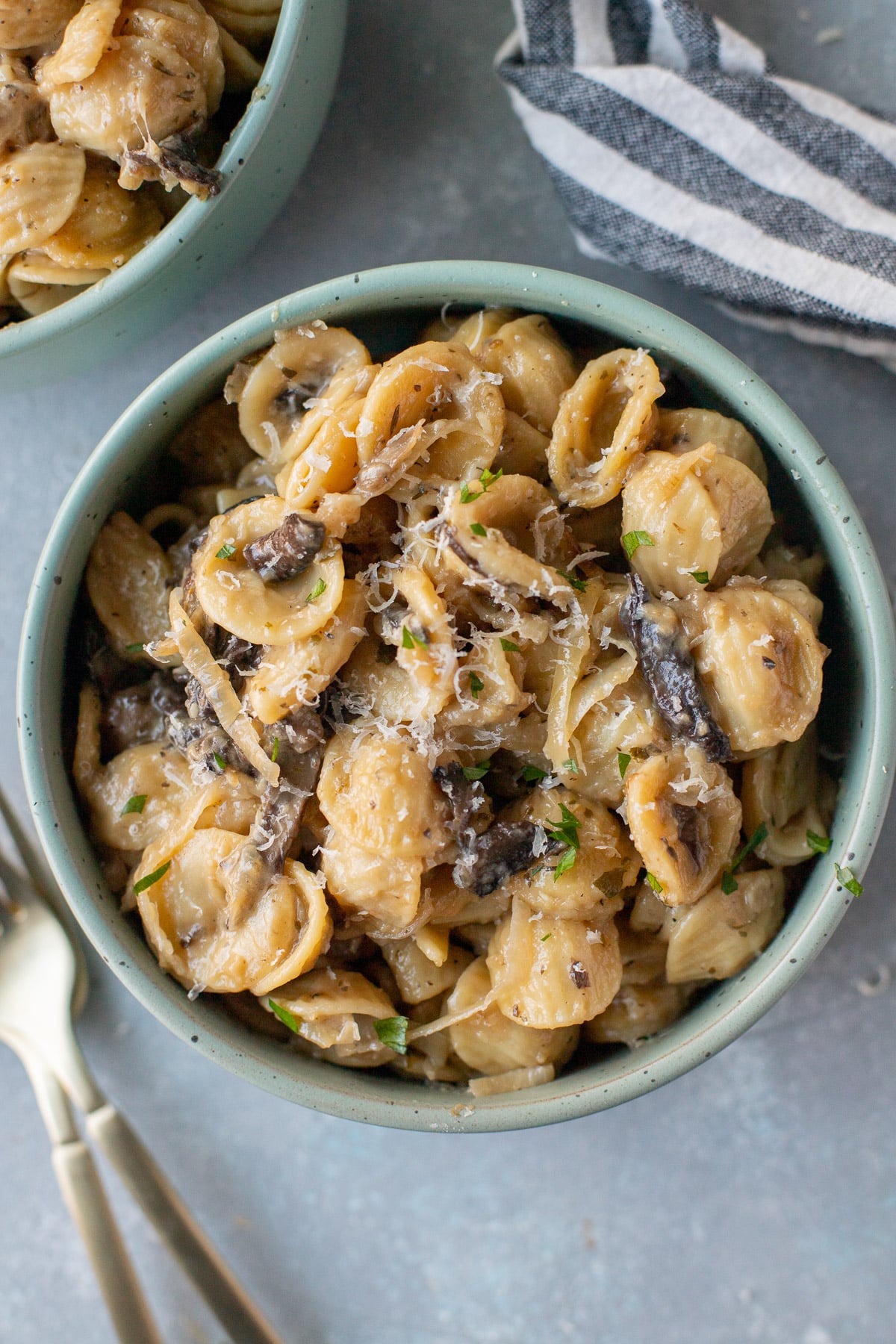 An overhead shot of a blue bowl of french onion soup pasta with another bowl, a towel and spoons on the side on a grey background