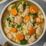 An overhead shot of Italian wedding soup in a white bowl with gold spoons and a bowl on the side on a grey background