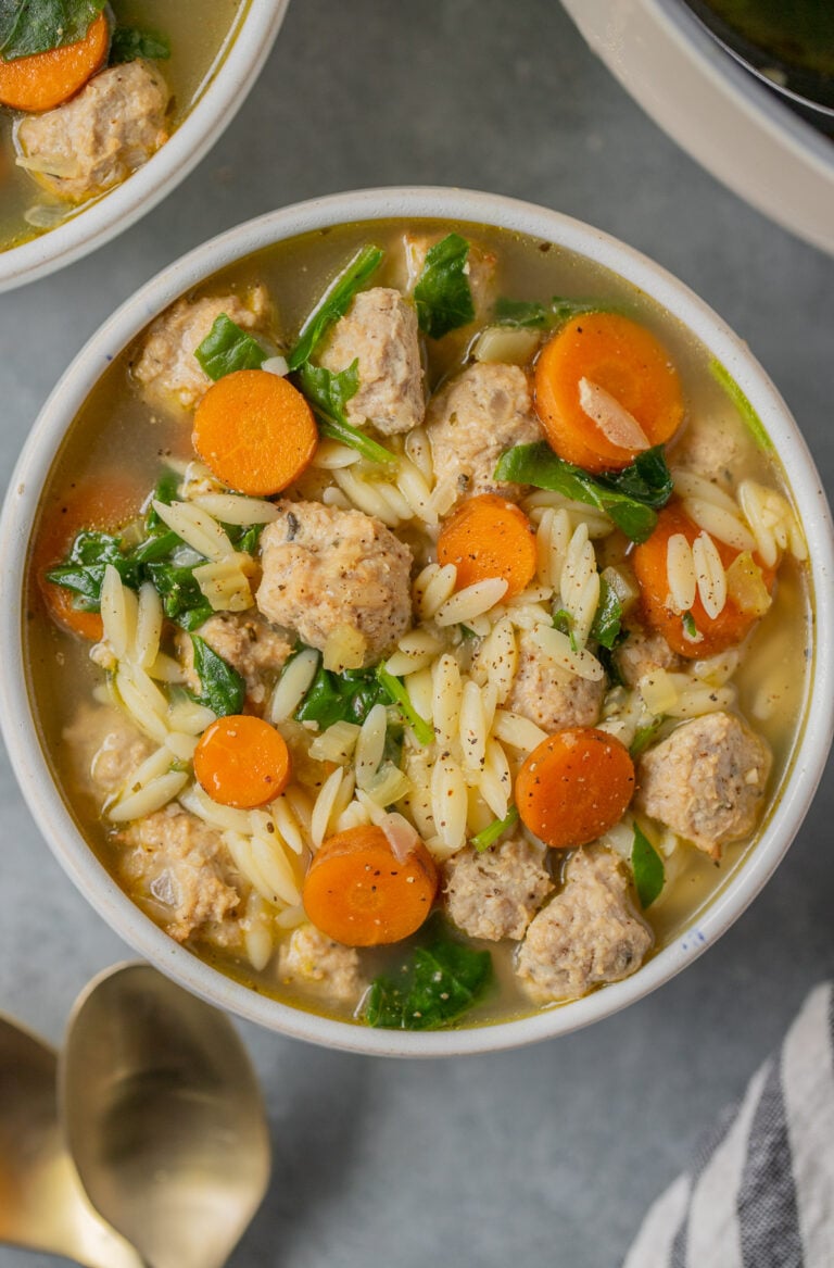 An overhead shot of Italian wedding soup in a white bowl with gold spoons and a bowl on the side on a grey background