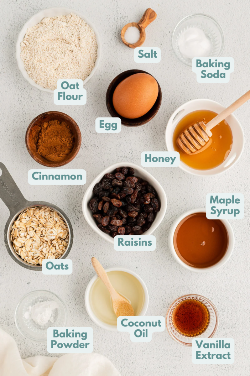 Ingredients for oatmeal raisin cookies in white bowls: raisins, rolled oats, oat flour, baking soda, powder, salt, cinnamon, coconut oil, maple syrup, honey, eggs, vanilla extract
