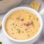 A white bowl of potato leek soup topped with bacon bits and cheese on a grey background with a dutch oven on the side