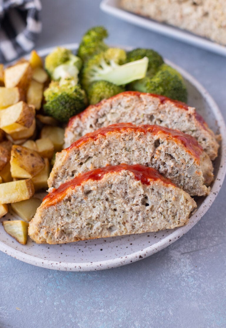 A plate with ground turkey meatloaf sliced beside some broccoli and roasted potatoes on a grey background