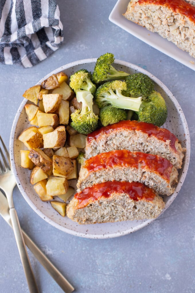 An overhead shot of a plate with ground turkey meatloaf sliced beside some broccoli and roasted potatoes on a grey background with forks on the side