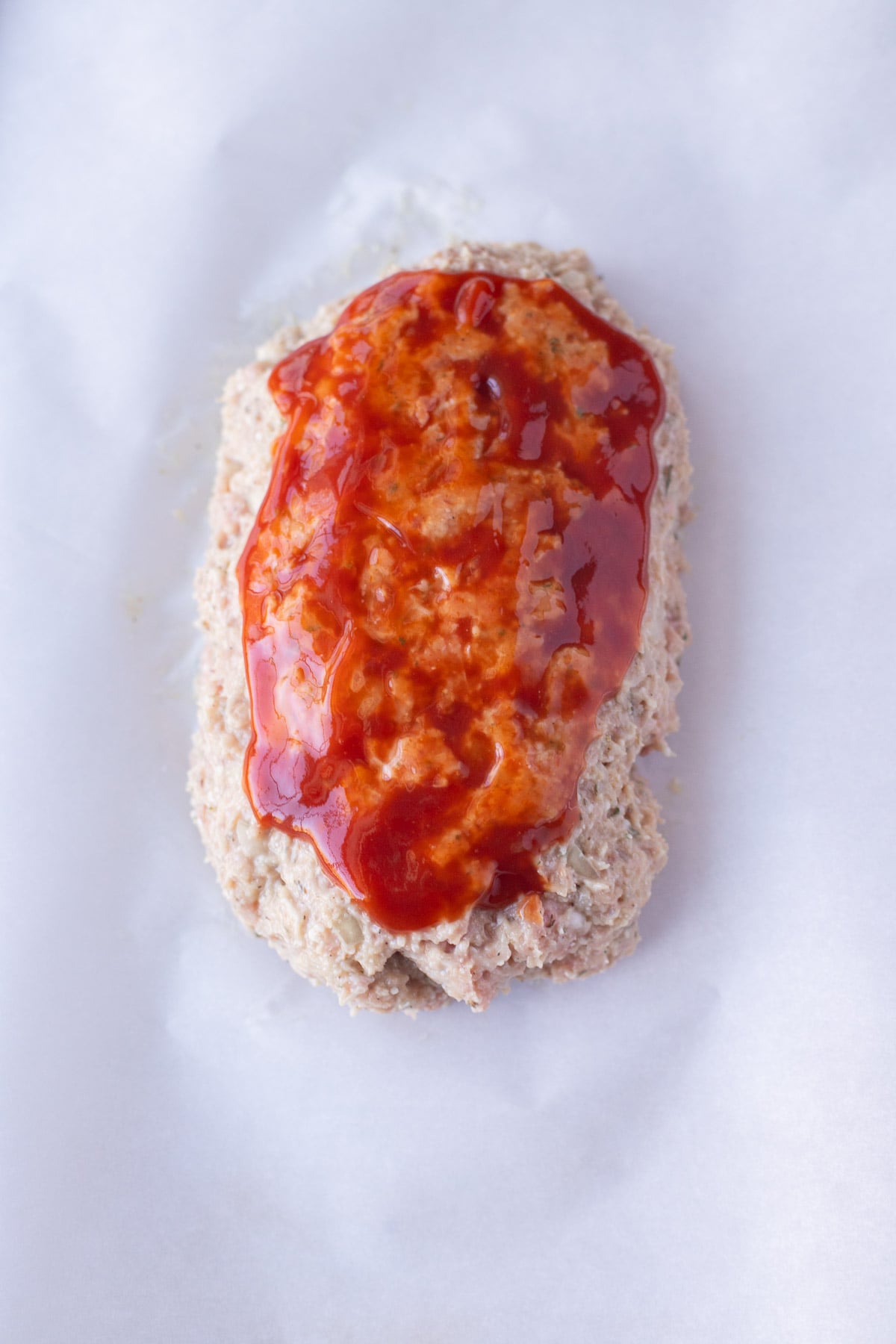 Raw ground turkey meatloaf topped with sauce on a piece of parchment paper