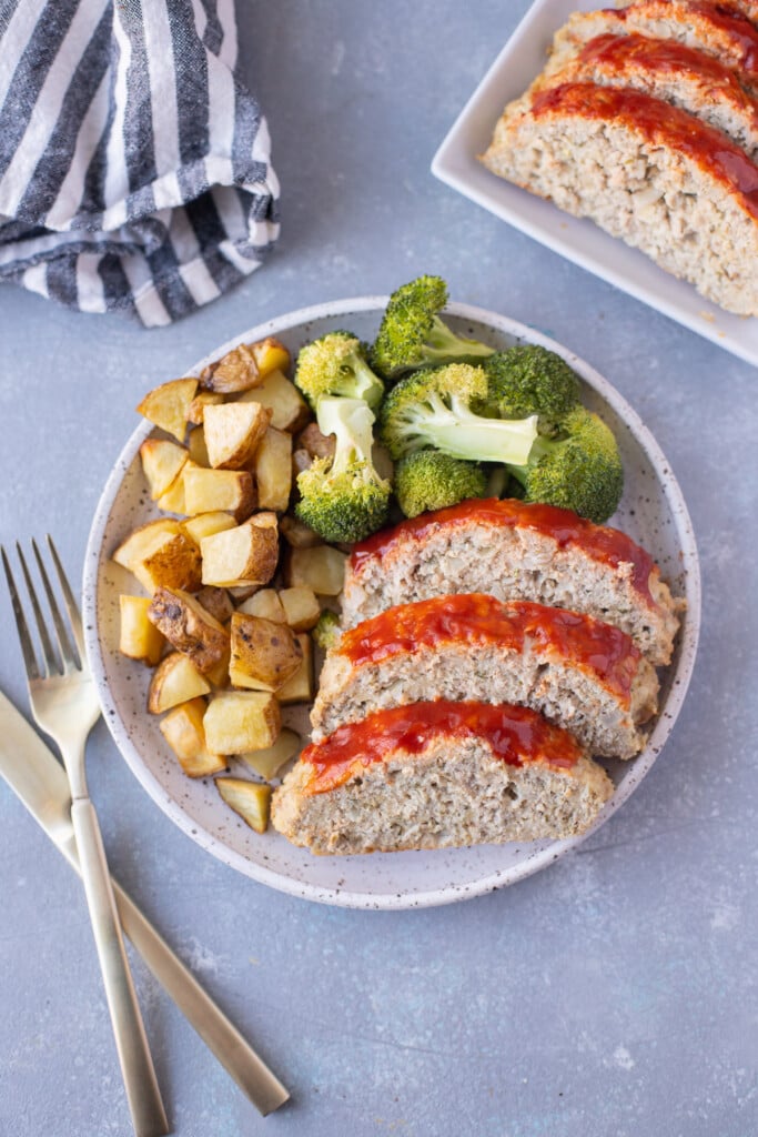 An overhead shot of a plate with ground turkey meatloaf sliced beside some broccoli and roasted potatoes on a grey background with forks and a plate of more meatloaf on the side