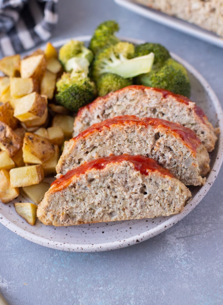 A plate with ground turkey meatloaf sliced beside some broccoli and roasted potatoes on a grey background
