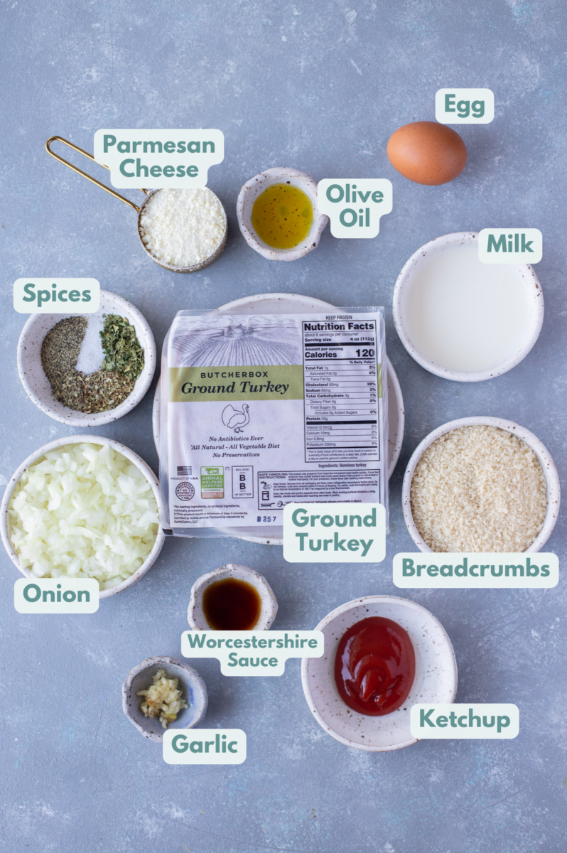Ingredients for healthy turkey meatloaf in white bowls: ground turkey, onion, garlic, bread crumbs, dried basil, dried parsley, dried oregano, salt, pepper, parmesan cheese, milk, egg
