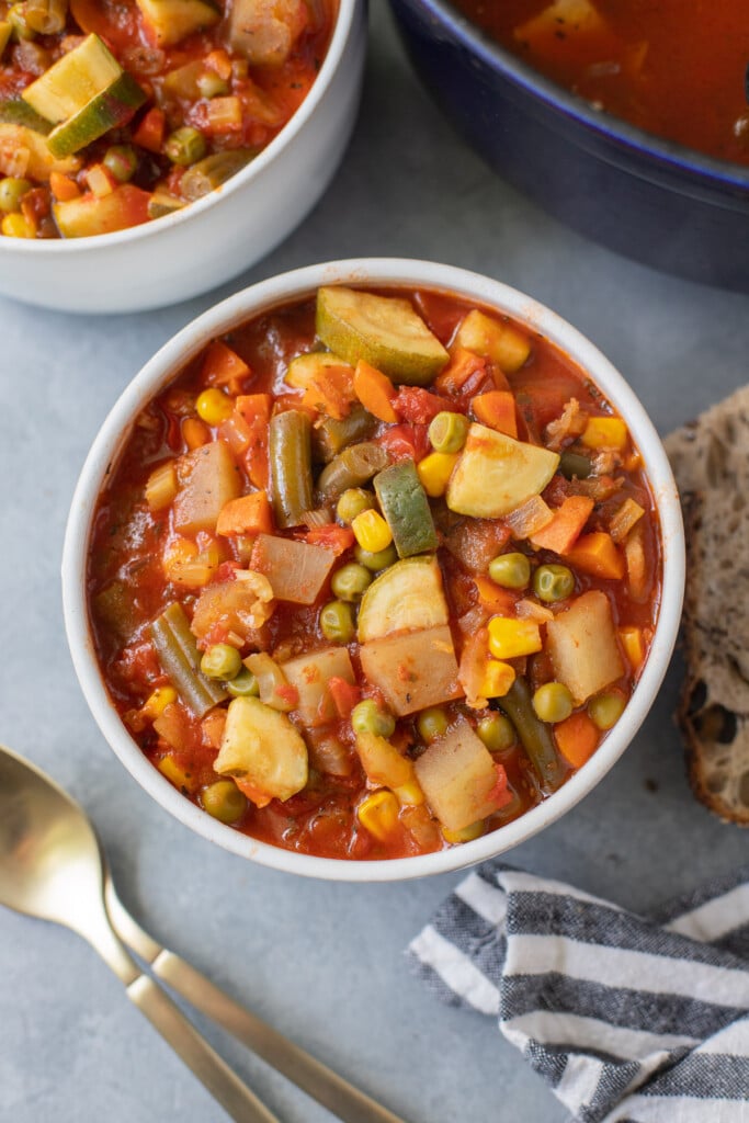 An overhead shot of a bowl of vegetable soup with gold spoons and crusty bread and a bowl and pot of soup on the side on a grey background