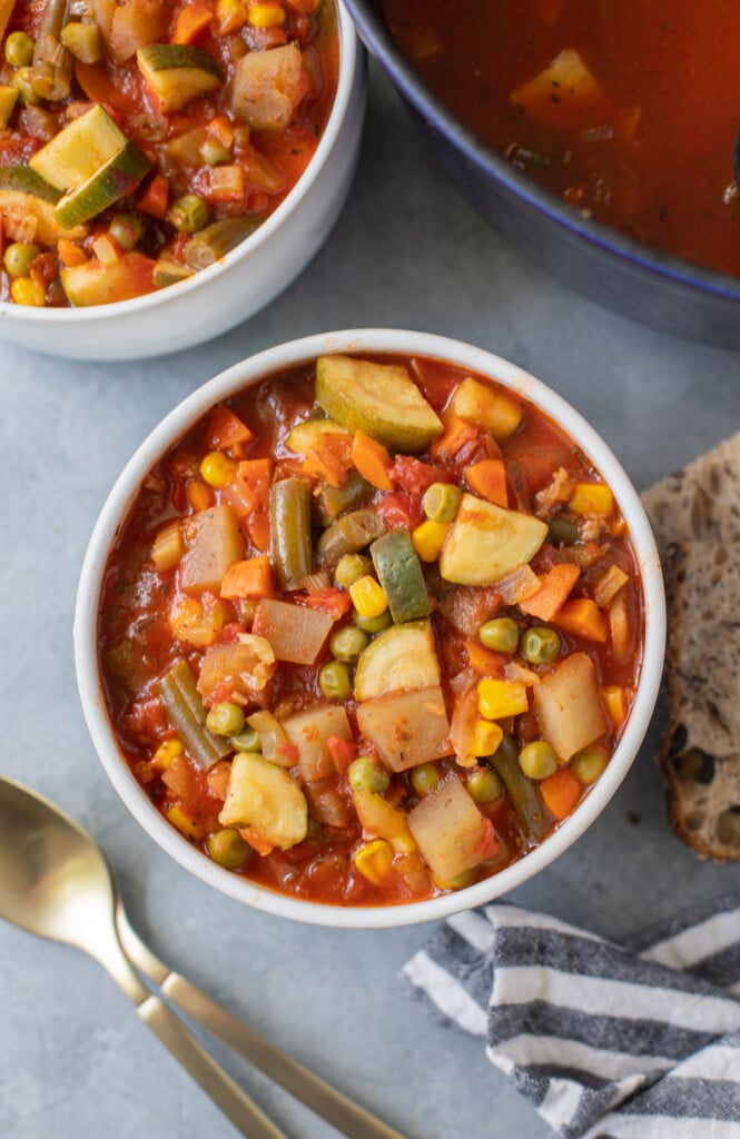 An overhead shot of a bowl of vegetable soup with gold spoons and crusty bread and a bowl and pot of soup on the side on a grey background