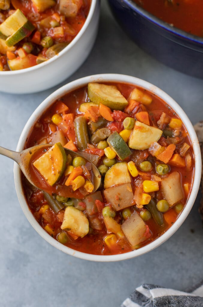 A white bowl of vegetable soup with a gold spoon taking a bite out of it with a bowl of soup in the background