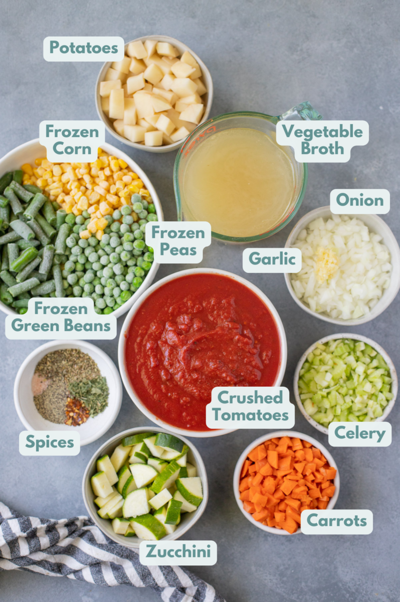 Ingredients for vegetable soup in white bowls: olive oil, onion, garlic, carrots, celery, potatoes, dried oregano, dried parsley, dried basil, salt, pepper, red pepper flakes, canned crushed tomato, broth, water, frozen corn, frozen green beans, peas, zucchini