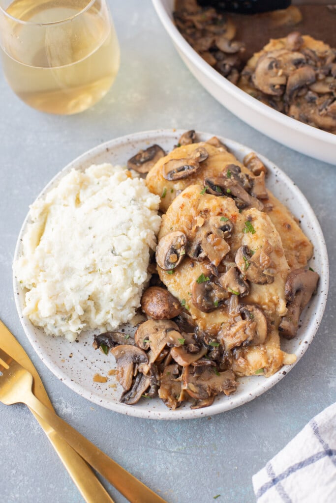 A white plate with Marsala Chicken and white rice with gold forks and a pan of chicken on the side