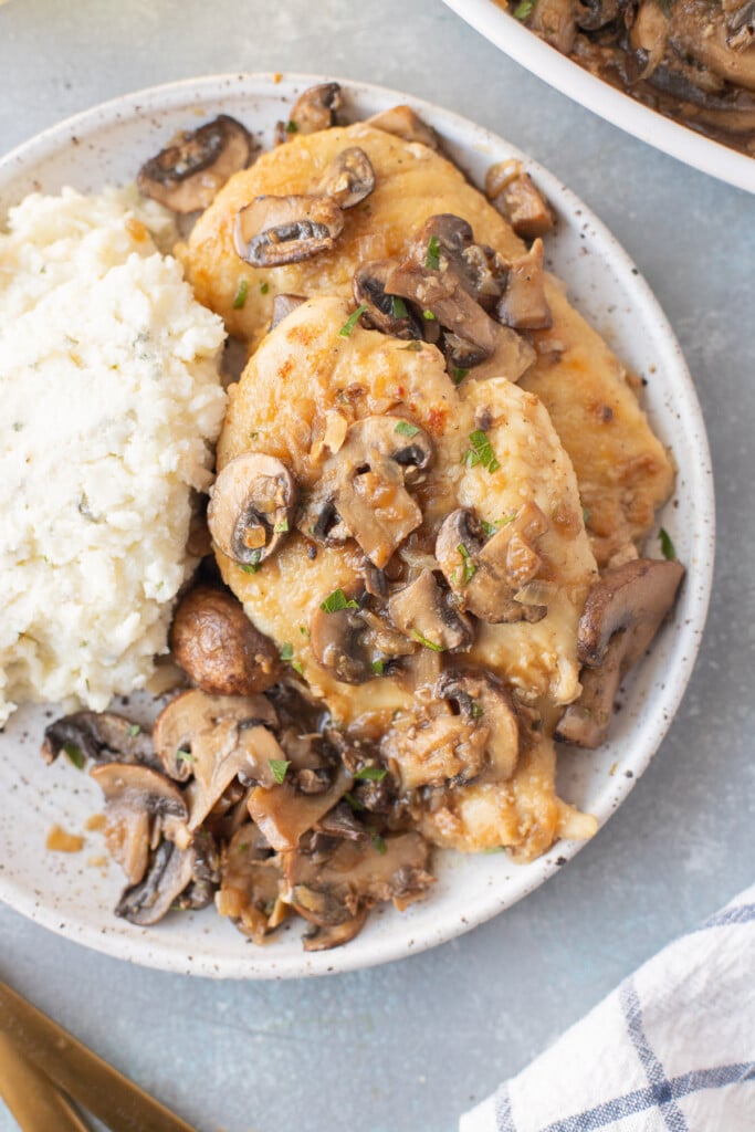 A close up on a white plate of chicken marsala with mashed potatoes and a pan of chicken in the background
