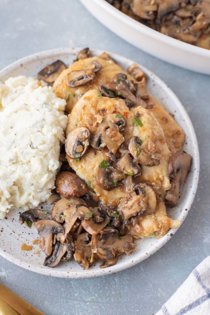 A close up on a white plate of chicken marsala with mashed potatoes and a pan of chicken in the background