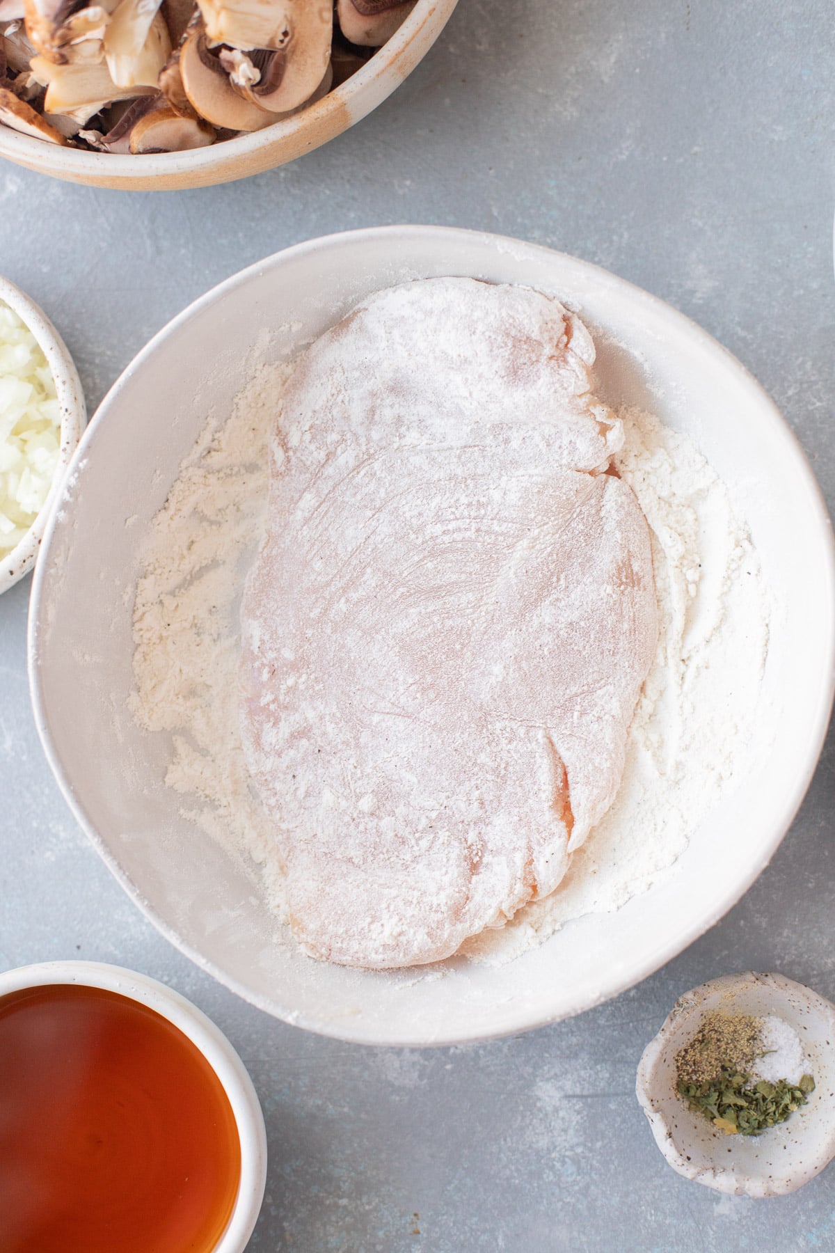 An overhead shot of a white shallow dish of flour and spices with a chicken breast getting dipped in it on a grey background