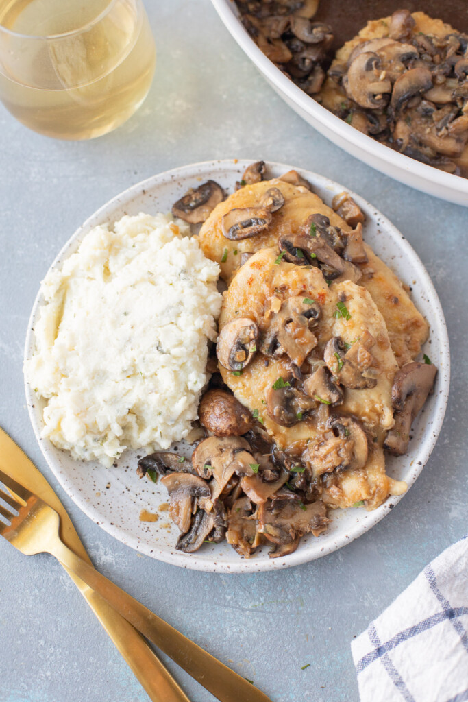 A white plate with Marsala Chicken and mashed potatoes with gold forks and a pan of chicken on the side