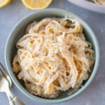 An overhead shot of a blue bowl of creamy lemon pasta with forks, a pan of pasta and lemons on the side
