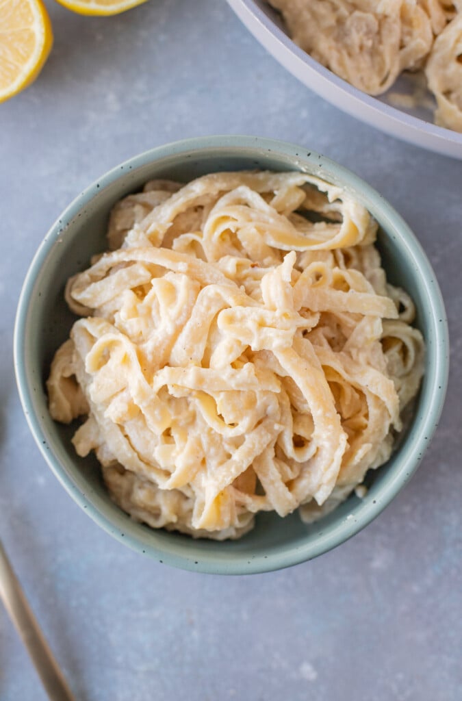 An overhead shot of a blue bowl of creamy lemon pasta with a pan of pasta on the side on a grey background