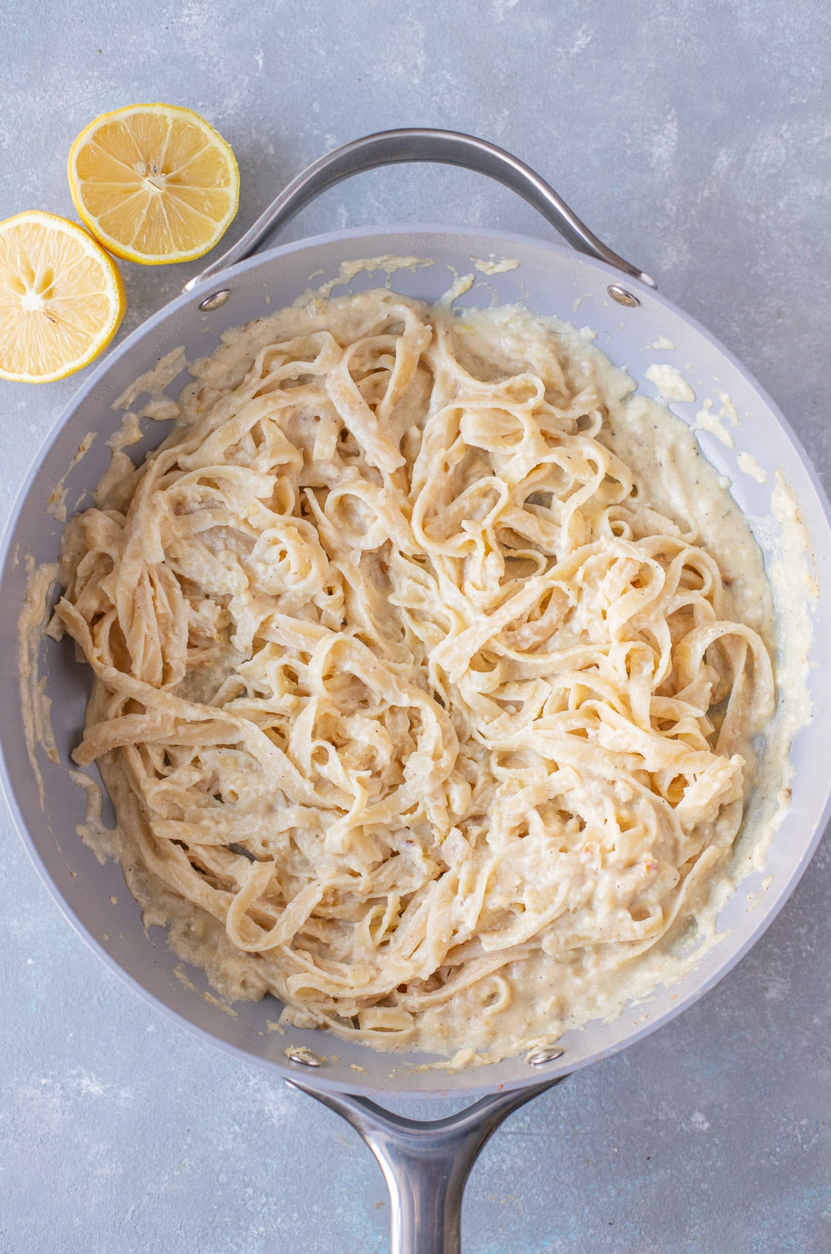 An overhead shot of a pan of creamy lemon pasta on a grey background with lemons on the side