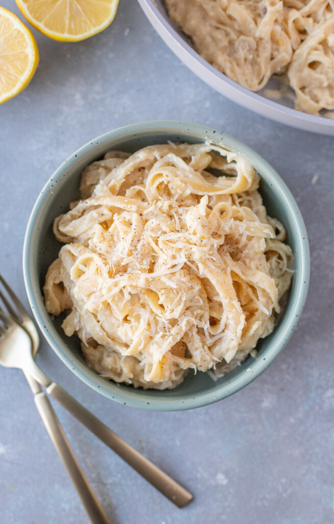 An overhead shot of a blue bowl of creamy lemon pasta with forks, a pan of pasta and lemons on the side on a grey background
