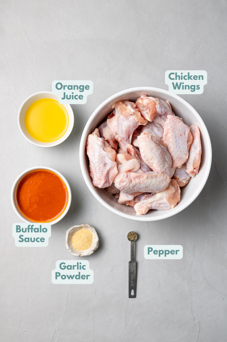 Ingredients for Crock pot Chicken Wings in small white bowls on a grey background: chicken wings, buffalo sauce, orange juice, garlic powder and pepper