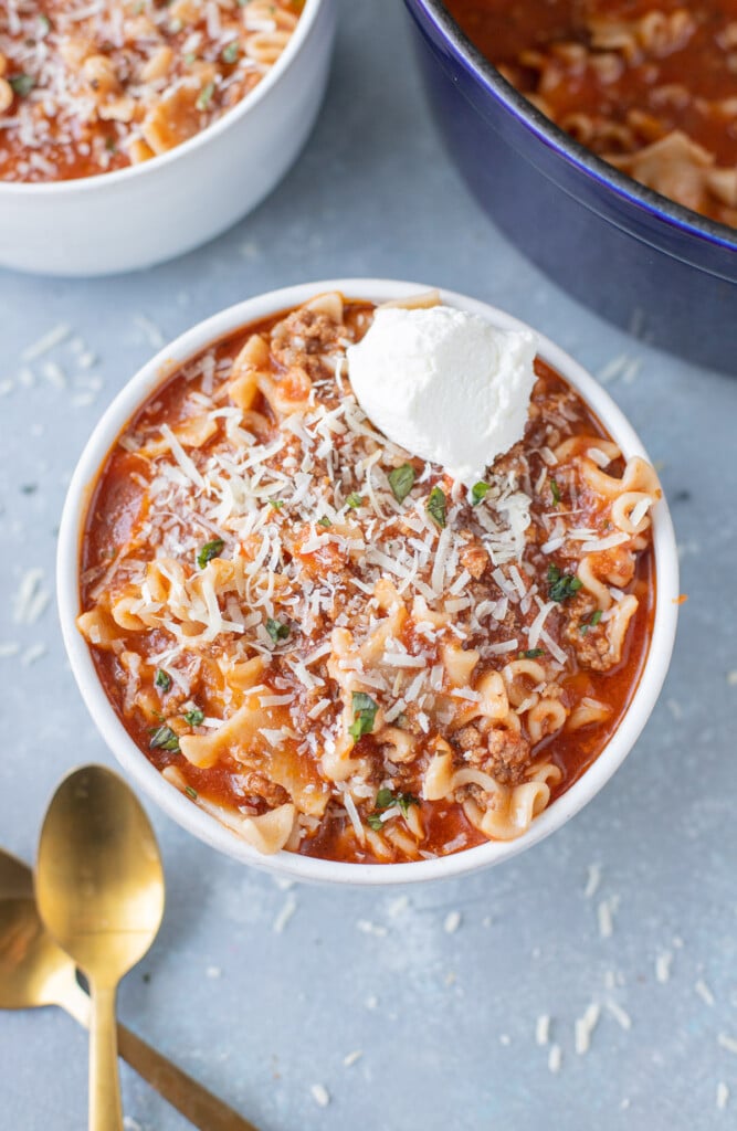 An overhead shot of lasagna soup with ricotta cheese on top with another bowl and pot on the side on a grey background