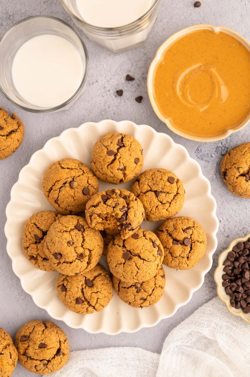 Protein cookies on a white plate with glasses of milk and nut butter in a bowl on the side.