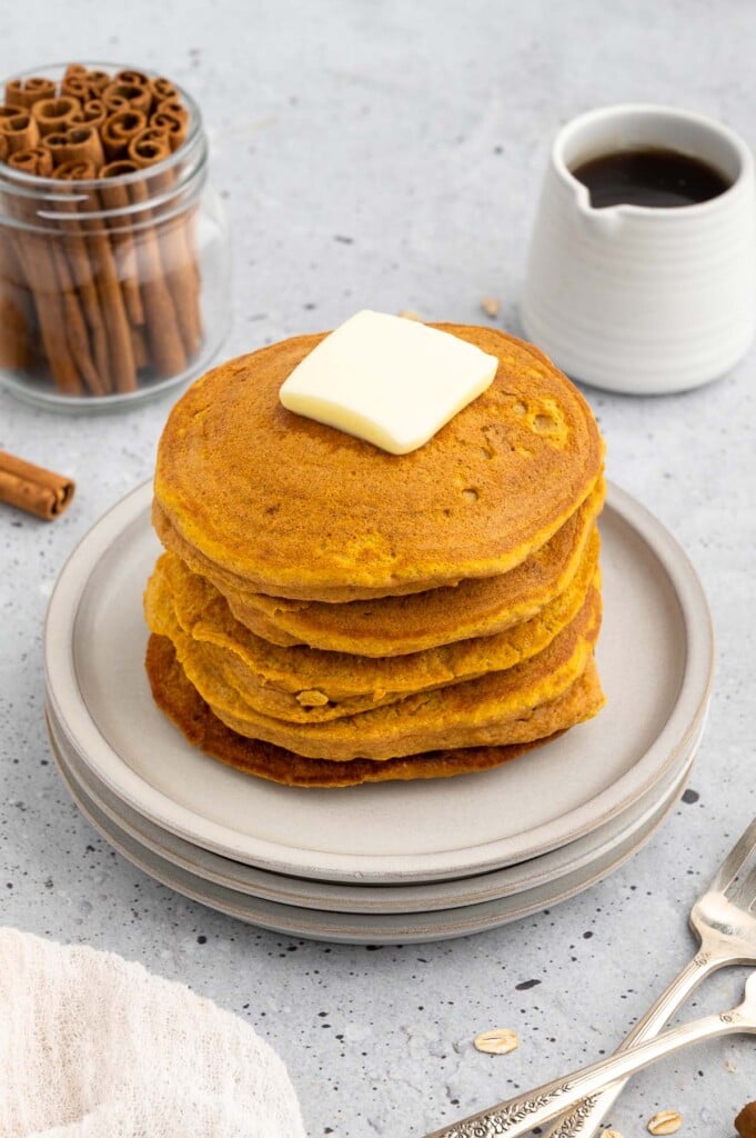 A stack of sweet potato pancakes on a white plate topped with a pat of butter with cinnamon sticks on the side on a grey background
