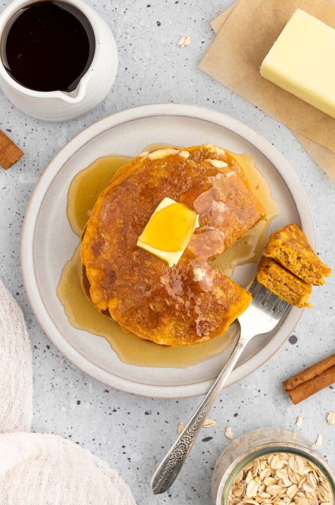 An overhead shot of a plate of sweet potato pancakes with a pat of butter and maple syrup on top and a fork with a bite on it and a dish of maple syrup on the side on a grey background
