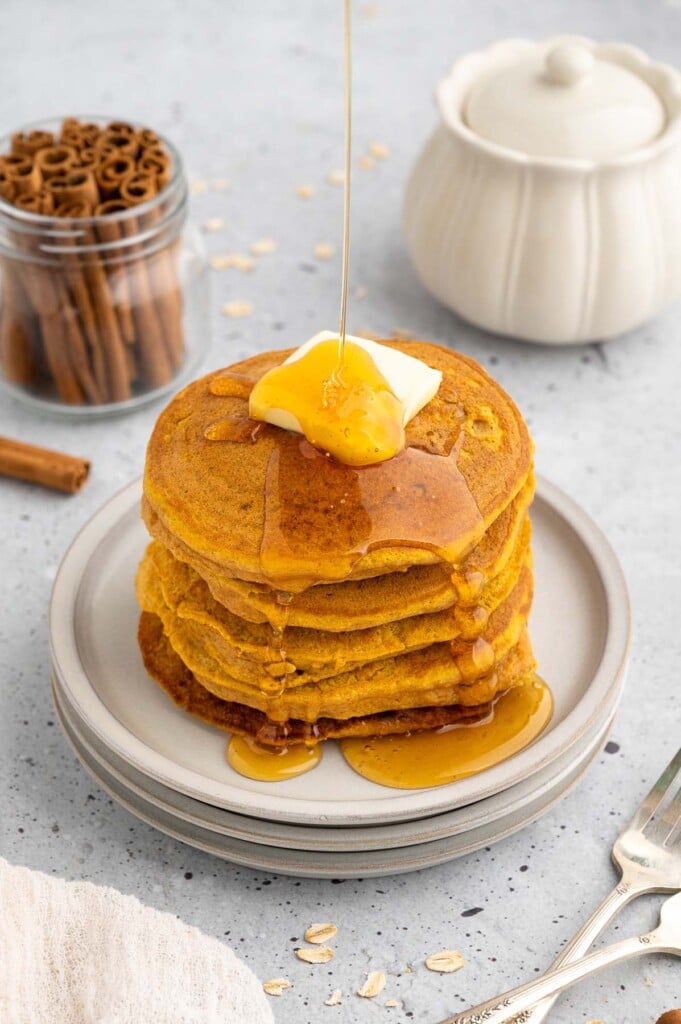 A stack of sweet potato pancakes on a white plate topped with a pat of butter with maple syrup being poured on top with cinnamon sticks on the side on a grey background